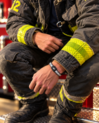 A close-up lifestyle shot of a firefighter in dark gray turnout gear with bright yellow reflective stripes, sitting on a fire truck. The firefighter is wearing a Black and White AV Edge Wristband