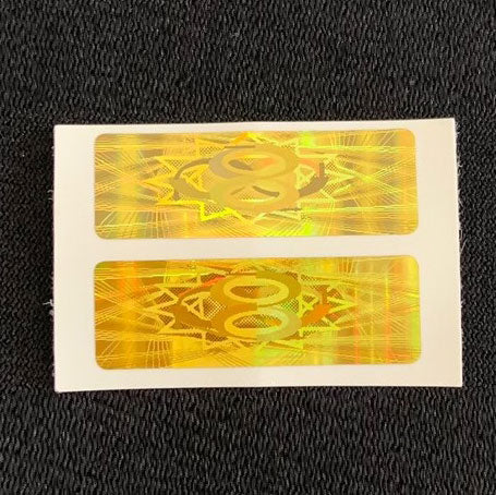 A close-up shot of two rectangular, golden Cell Phone EMF Protect Filters on their adhesive backing. The holograms feature a geometric star and circle pattern, highlighting the EF Technology used as a frequency-based filter to provide biological EMF protection for mobile devices.